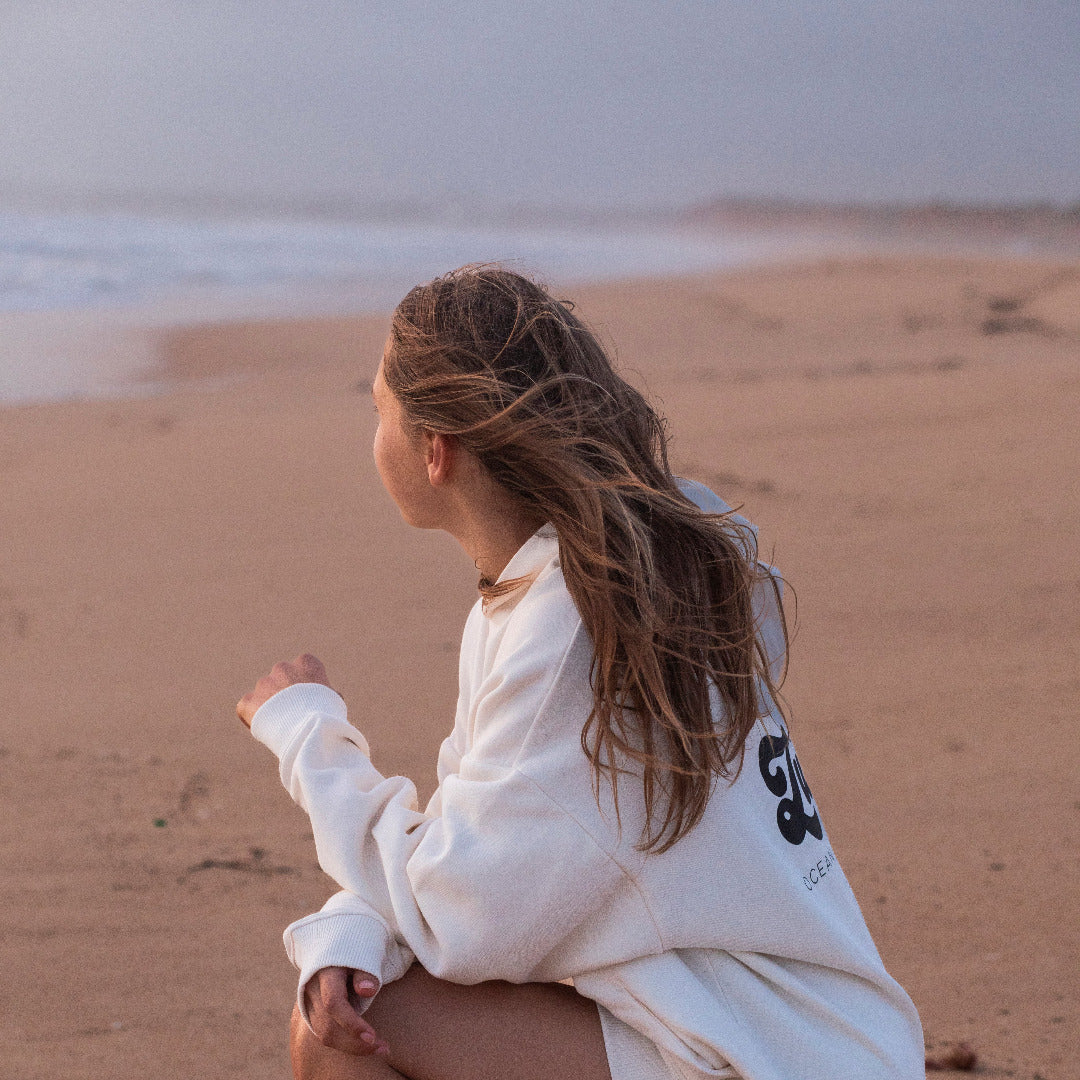 Eine junge Frau sitzt am Strand und schaut aufs Meer, sie trägt einen weissen Hoodie von Zurfday