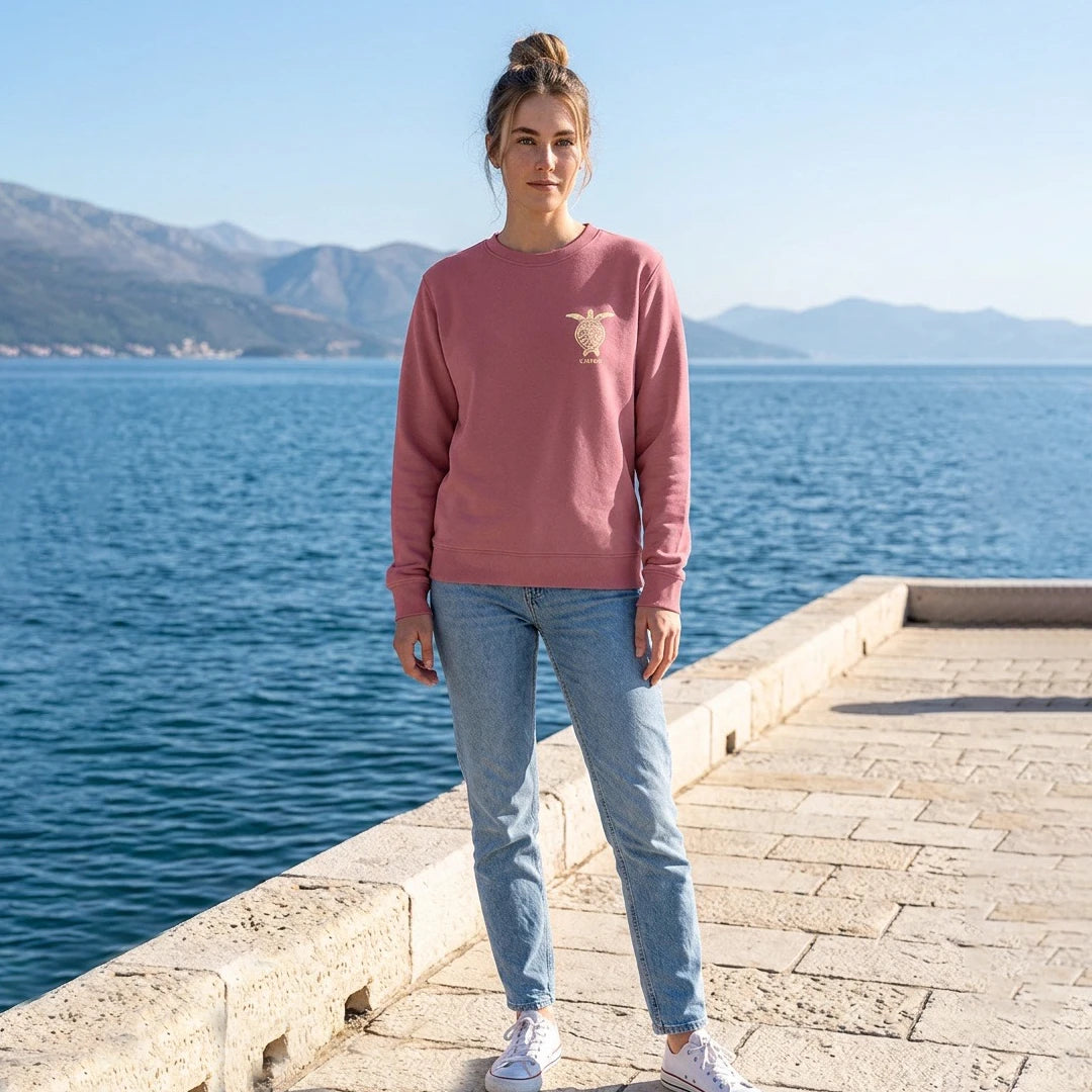 Eine junge Frau trägt den Carry the Ocean Sweater in Antique Rosa mit großem Schildkröten-Print an der Küste.