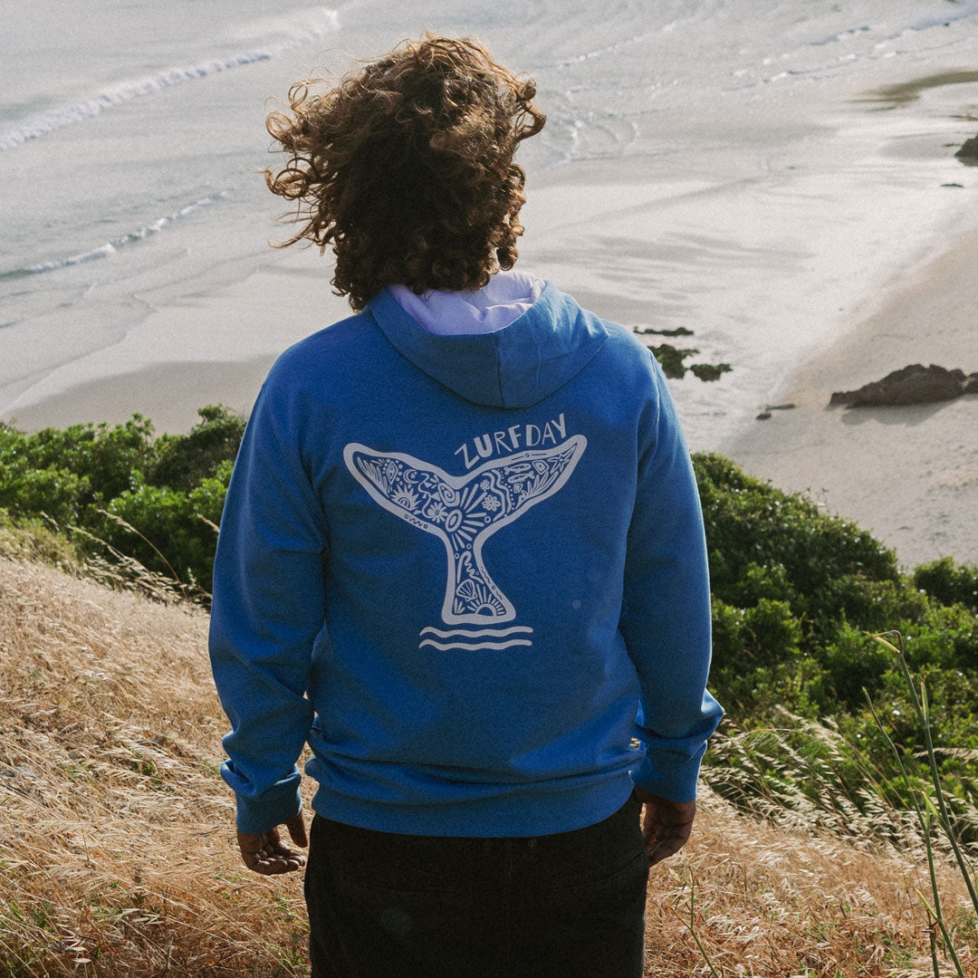 Surfer steht am Strand und schaut aufs Meer, er trägt einen Hoodie von Zurfday