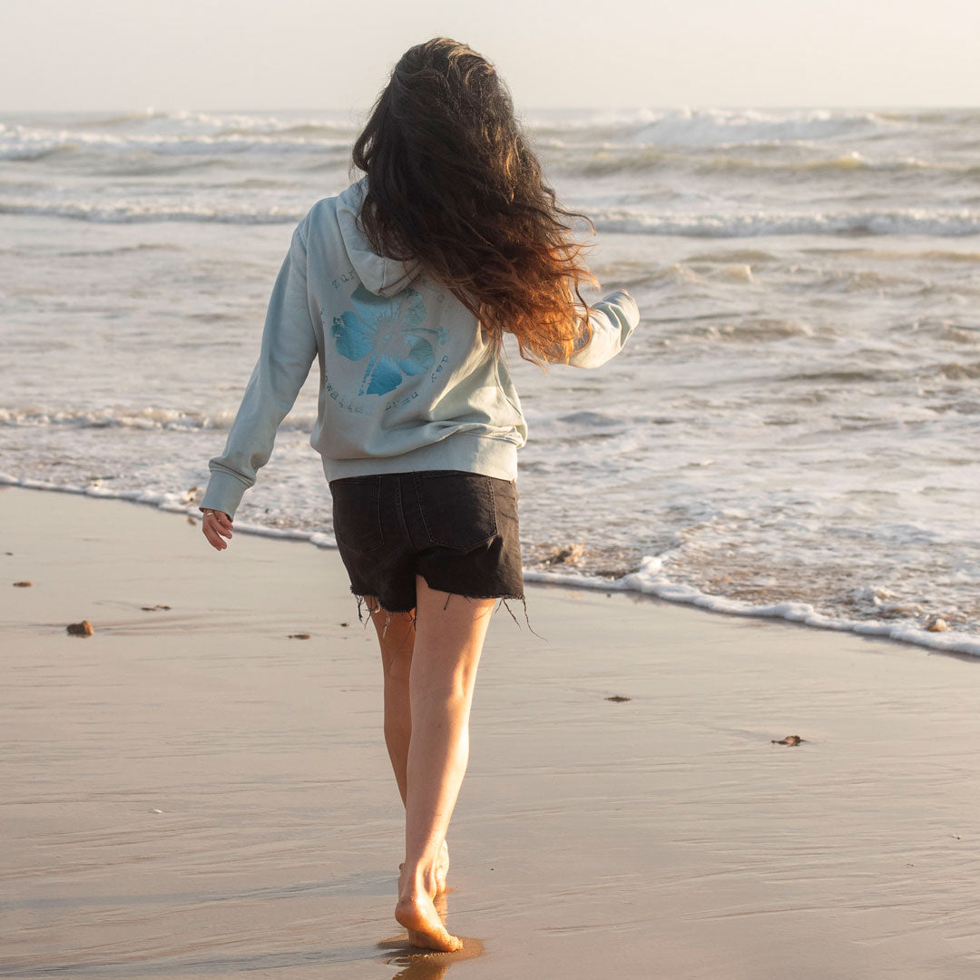 Eine junge Frau läuft zum Meer, sie trägt einen grünen Hoodie von Zurfday mit einer großen Hawaii Blume
