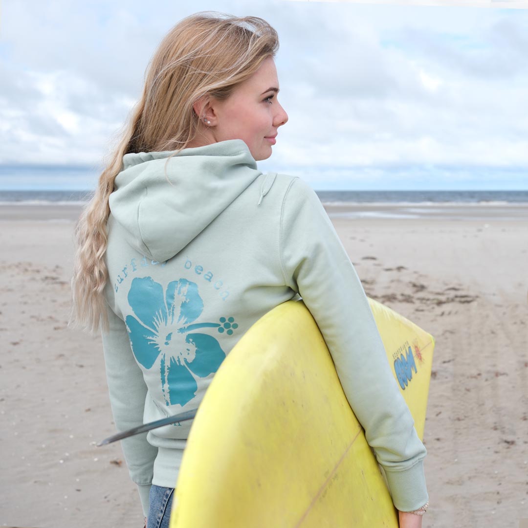 Junge Frau mit Surfbrett unterm Arm trägt den Zurfday Hoodie Haiwaiian Luau.