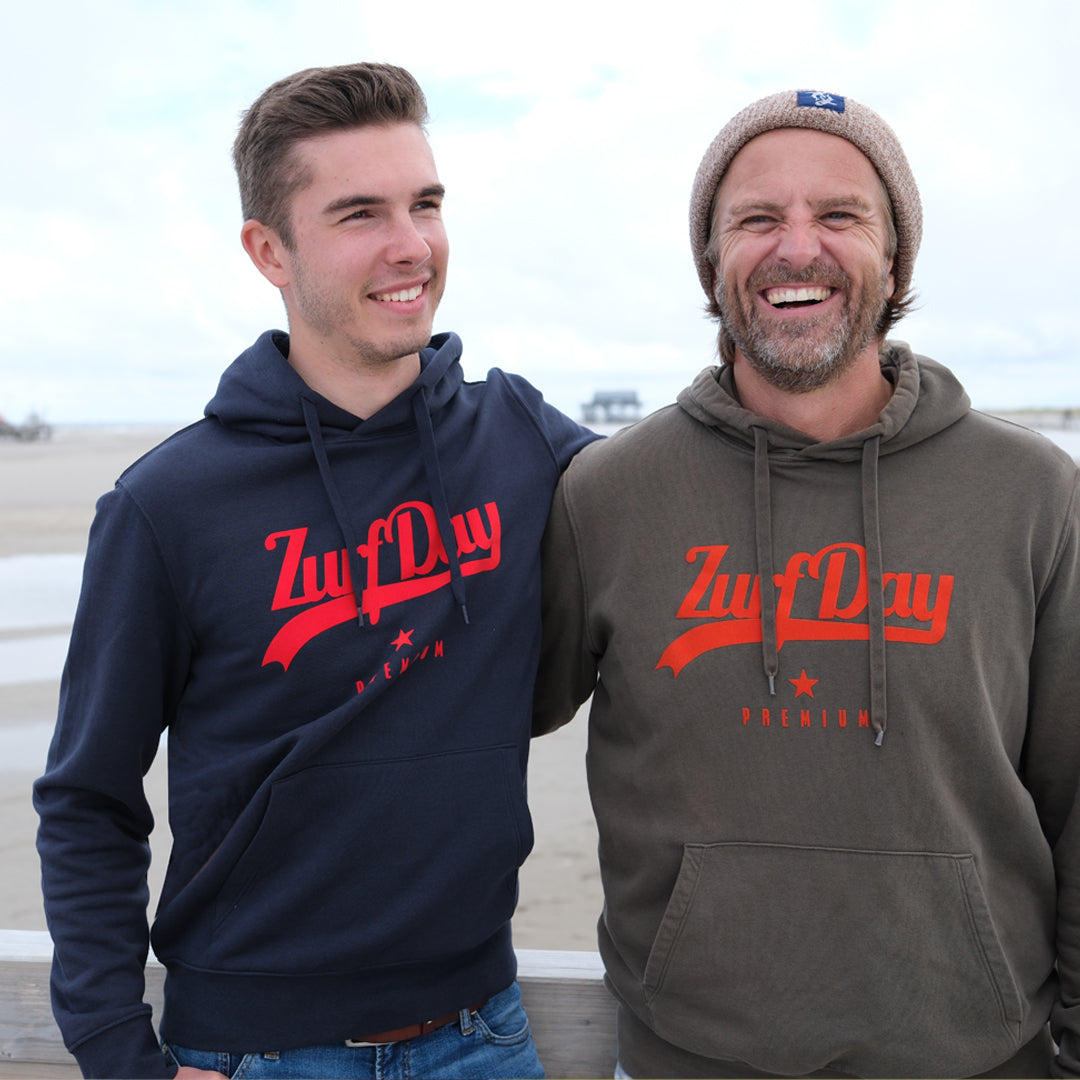 Zwei Freunde stehen am Strand. sie lachen in die Kamera. Beide tragen ein Hoodie von Zurfday, einen in Navi Blau mit rotem Print und einen  Hoodie in Olive mit orangefarbener Schrift