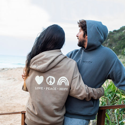 Ein junges Surfr Paar am Strand, beide tragen den Love Peace Hope Hoodie von Zurfday