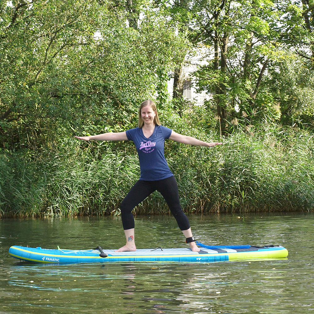 Junge blonde Frau macht Yoga auf dem Sup-Board und trägt ein blaues Zurfday T-Shirt mit pinkem glitzer Aufdruck