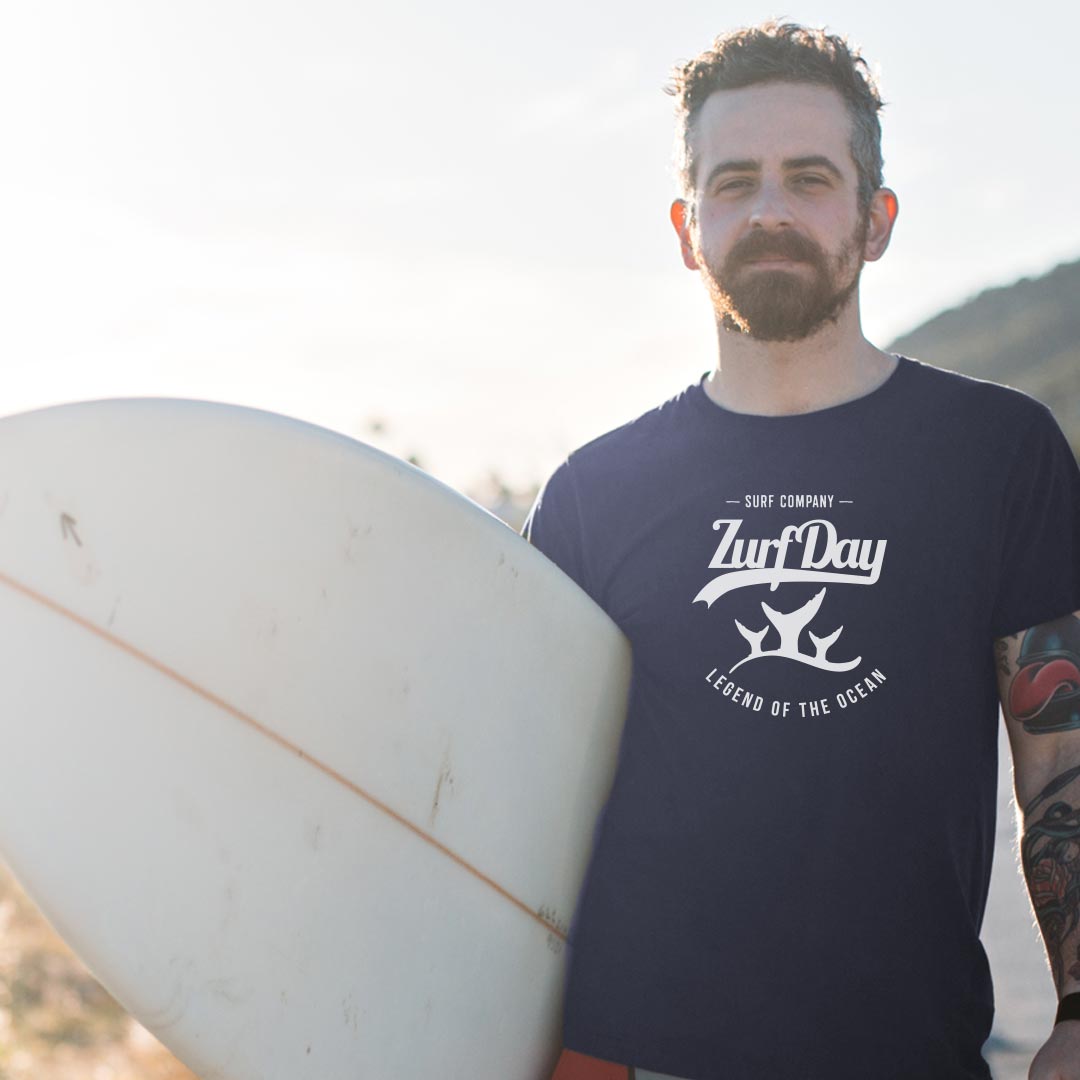 Man trägt dunkles Zurfday-T-Shirt mit Print und hat Surfbrett unter dem Arm.