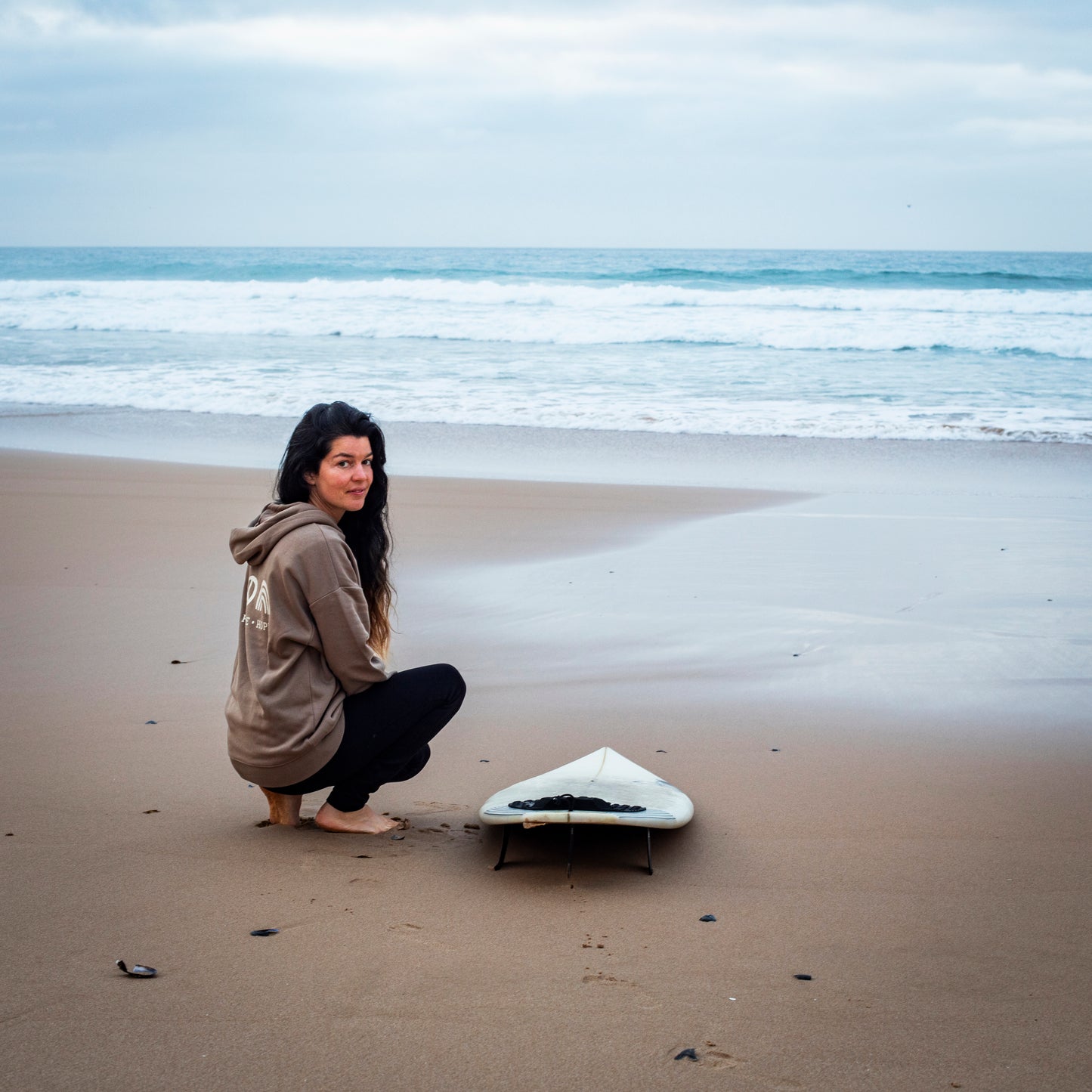 Eine junge Surferin kniet neben dem Board und blickt in die Kamera, sie trägt einen Zurfday Hoodie
