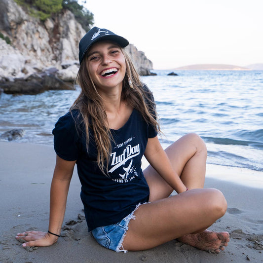 Junge Frau sitzt lachend am Strand, sie trägt ein blaues Zurfday T-Shirt