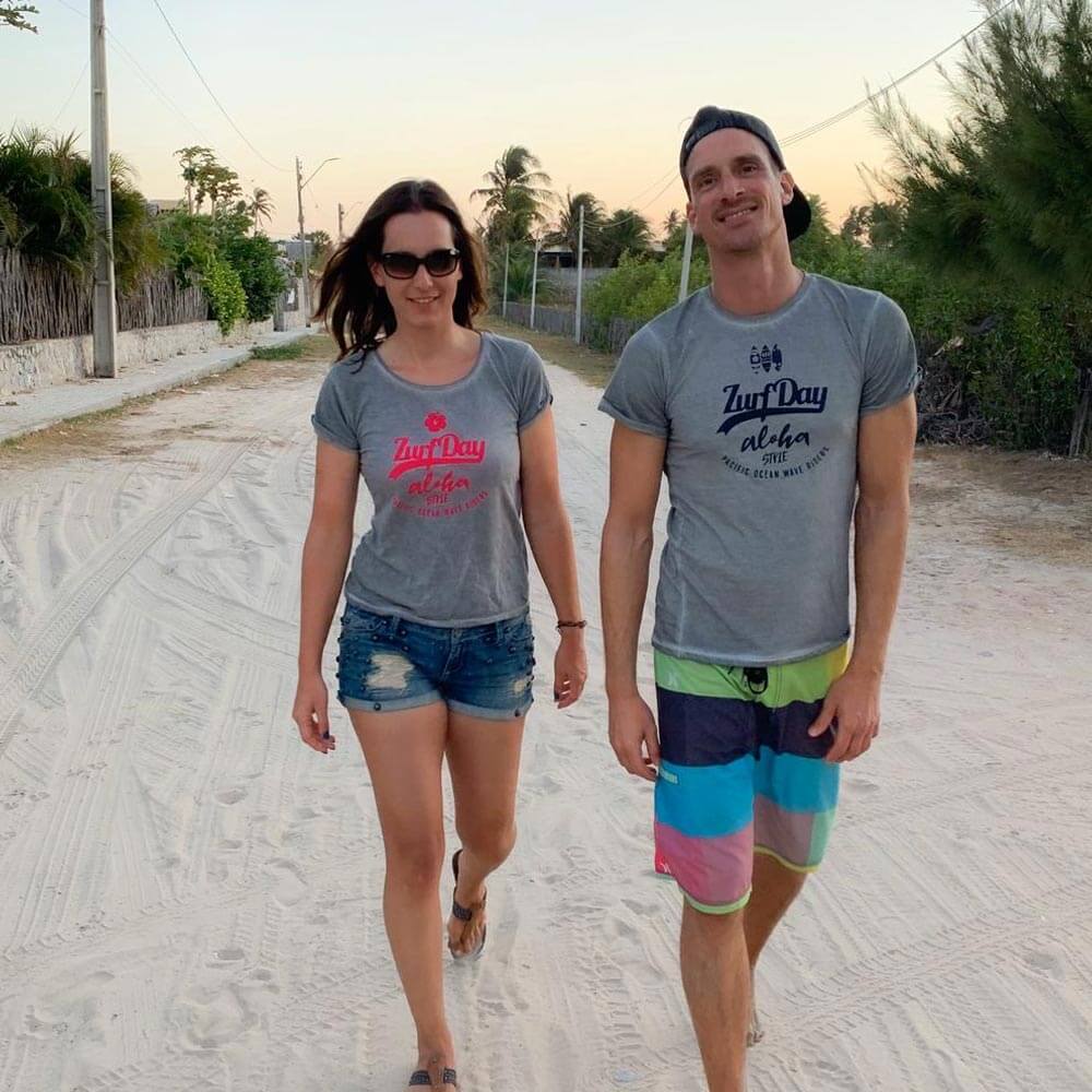 Ein glückliches Paar geht am Strand spazieren und beide tragen das bedruckte Aloha T-Shirt von Zurfday